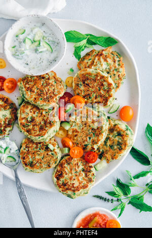 Zucchini Krapfen auf einer Keramikplatte mit Trauben Tomaten und Joghurt dip auf der Seite von der Ansicht von oben fotografiert. Stockfoto