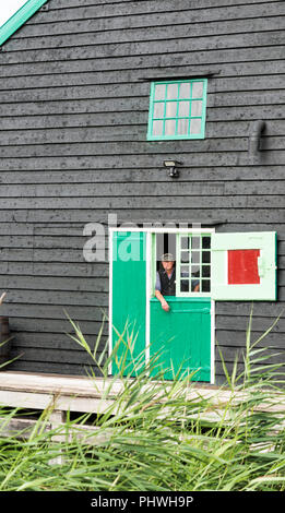 Zaandam, Niederlande, 28-Aug-2018: Niederländische Bauer mit Hut auf und ein Rohr in den Mund schaut aus einem alten Bauernhof mit Fenstern mit Fensterläden, Zaandam ist berühmt für seine alten Bauernhöfen und Leben, wie es war vor hundert Jahren Stockfoto