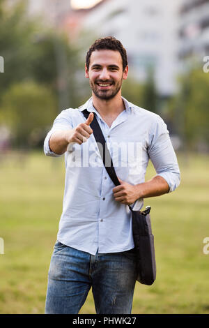Junge Unternehmer steht im Park und zeigt den Daumen nach oben. Stockfoto