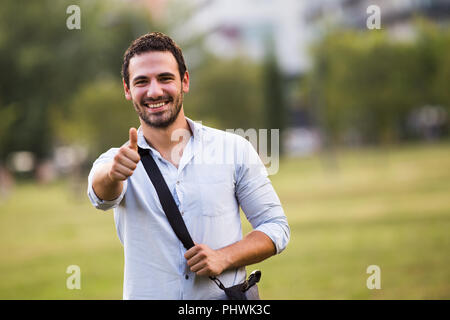 Junge Unternehmer steht im Park und zeigt den Daumen nach oben. Stockfoto