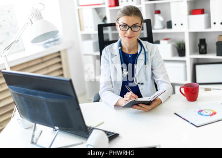 Ein junges Mädchen in einem weißen Gewand sitzt an einem Tisch im Büro und hält einen Stift und ein Notebook in der Hand. Ein Stethoskop um den Hals hängt. Stockfoto