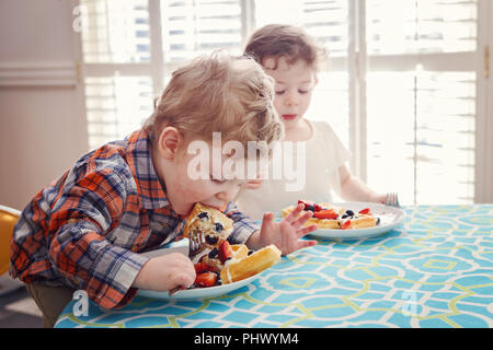 Zwei glückliche Kinder Zwillinge junge Mädchen essen Frühstück Waffeln mit Früchten am Tisch sitzen im sonnigen Küche am frühen Morgen mit Sonnenlicht von hinten Stockfoto