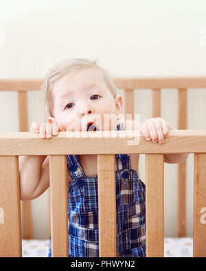 Porträt einer cute adorable lustig Baby neun Monate im Bett stehend Krippe kauen Essen saugen hölzernen Seiten, Blick in die Kamera, Lifestyle everyda Stockfoto