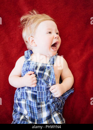 Portrait von cute adorable Kaukasischen smiling baby junge Mädchen liegt auf dem Boden rote Decke im Kinderzimmer schreiend weinend Weg schauen, natürliche Fenster Licht, Stockfoto