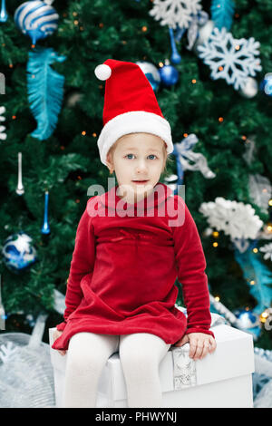 Portrait von Glücklich lächelnde lustig Kaukasier Kind girl Toddler mit blauen Augen im roten Kleid und Santa Claus hat sitzen auf Gist, indem neue Jahr Baum, lif Stockfoto