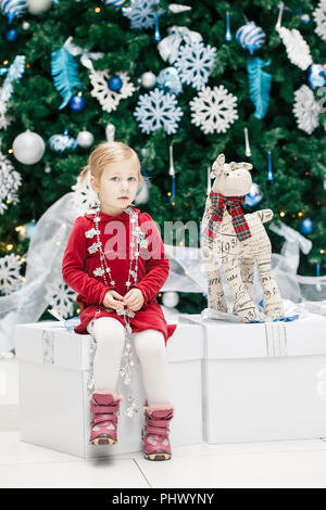 Portrait von Blond kaukasische Baby girl Toddler mit blauen Augen im roten Kleid sitzen durch neues Jahr Baum in der Nähe von Hirsch Spielzeug, emotionale lifestyle Bild. Stockfoto