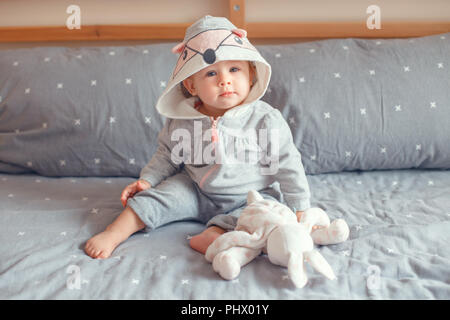 Portrait von cute adorable Kaukasischen blond Lächelnden Mädchen mit blauen Augen in grau Pyjama mit Fox Katze Tier Haube sitzen auf dem Bett im Schlafzimmer und Halten Stockfoto