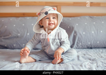 Portrait von cute adorable Kaukasischen blond Lächelnden Mädchen mit blauen Augen in grau Pyjama mit Fox Katze Tier Haube sitzen auf dem Bett im Schlafzimmer. Natürliche Stockfoto