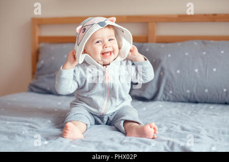 Portrait von cute adorable Kaukasischen blond Lächelnden Mädchen mit blauen Augen in grau Pyjama mit Fox Katze Tier Haube sitzen auf dem Bett im Schlafzimmer. Natürliche Stockfoto