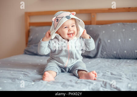 Portrait von cute adorable Kaukasischen blond Lächelnden Mädchen mit blauen Augen in grau Pyjama mit Fox Katze Tier Haube sitzen auf dem Bett im Schlafzimmer. Natürliche Stockfoto