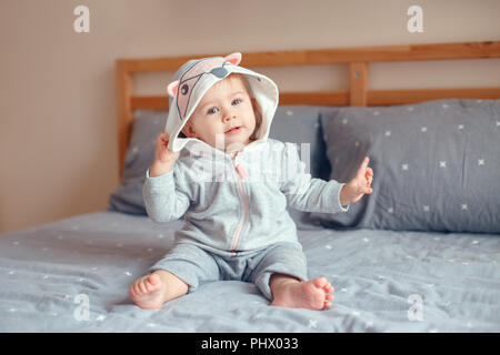 Portrait von cute adorable Kaukasischen blond Lächelnden Mädchen mit blauen Augen in grau Pyjama mit Fox Katze Tier Haube sitzen auf dem Bett im Schlafzimmer. Natürliche Stockfoto