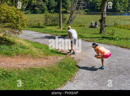 BLOWING ROCK, NC, USA -8/23/18: zwei Läufer, männlich und weiblich, vor der Ausführung um Bass Lake Stretching und zwei Frauen sitzen auf einer Bank in der Nähe des Sees. Stockfoto