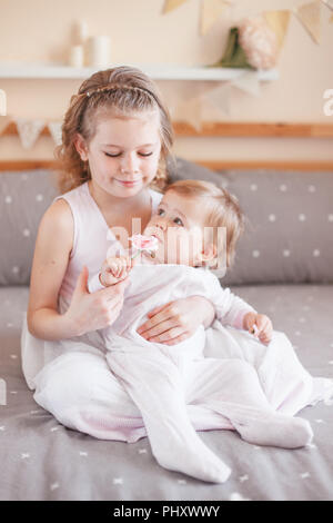 Portrait von cute adorable white Kaukasische Mädchen, dass kleine Schwester Baby auf runden Knien. Lächelnde Kinder sitzen zusammen auf dem Bett im Schlafzimmer suchen i Stockfoto