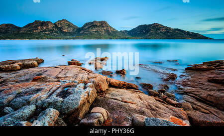 Die Gefährdungen in Freycinet Nationalpark in der Dämmerung, von Coles Bay in Tasmanien, Australien gesehen, mit orangefarbenen Flechten auf Felsen im Vordergrund. Stockfoto