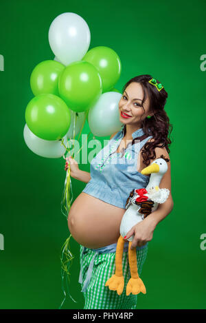Schwangere Frau mit Ballons und Storch auf einem grünen Hintergrund. Er schaut auf seinen Bauch, in Erwartung. Stockfoto