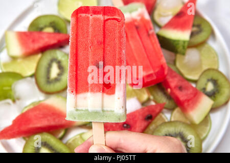 - Nahaufnahme gefrorenes Obst Beerensaft, hält eine weibliche Hand auf dem Hintergrund einer Platte mit Stücken von Wassermelone, Kiwi, Kalk und Eiswürfel Stockfoto