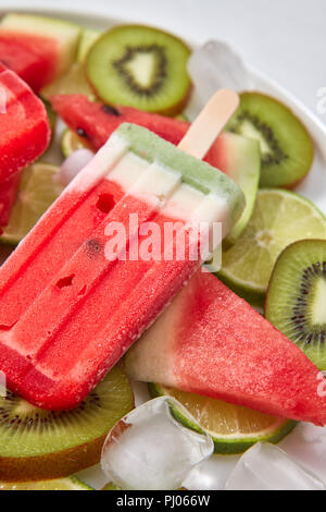 Close-up Obst frozen Smoothies auf einem Stick in Form eines Stück Wassermelone in einem Teller mit Eiswürfel und Früchte auf einem grauen Hintergrund. Eis am Stiel. Flach Stockfoto