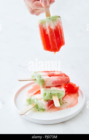 Eis am Stiel in einem Teller mit Spritzern. Hand hält eine Frau eines schmelzenden Eis auf einem weißen Hintergrund mit Kopie Raum Stockfoto