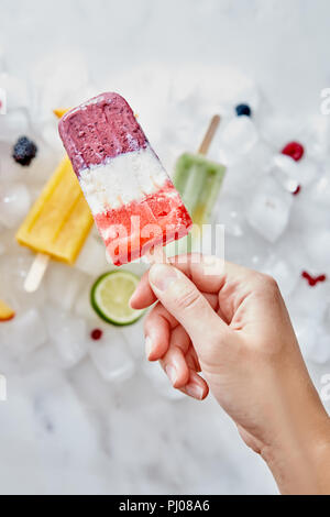 Hausgemachte frozen Smoothies auf einem Stock, die eine weibliche Hand vor dem Hintergrund der Eiswürfel mit verschiedenen Beeren und Eis. Stockfoto