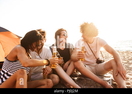 Foto der aufgeregten Gruppe von Freunden im Freien am Strand sitzen und Bier trinken Miteinander reden. Stockfoto