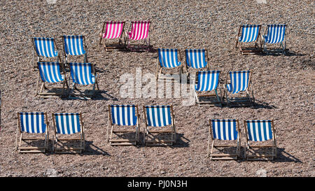 Liegestühle aufgereiht auf einem Kieselstrand am Meer. Es sind keine Personen auf dem Foto. Zwei der gestreiften Stühlen sind eine andere Farbe Stockfoto