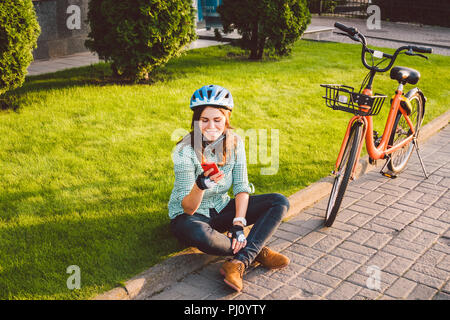 Mensch und Stadt rollenden Fahrrad, umweltfreundliche Verkehrsmittel. Schönen jungen kaukasischen Arbeiterin sitzen ruht auf dem Gras verwendet eine rote mobil Stockfoto