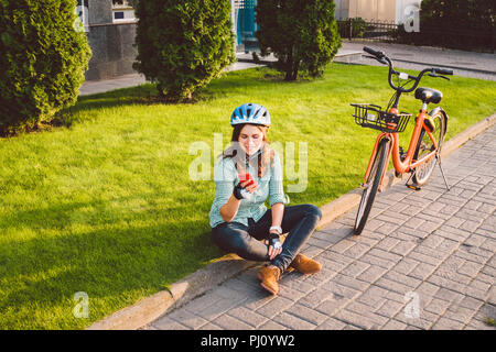 Mensch und Stadt rollenden Fahrrad, umweltfreundliche Verkehrsmittel. Schönen jungen kaukasischen Arbeiterin sitzen ruht auf dem Gras verwendet eine rote mobil Stockfoto