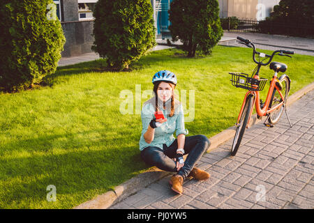 Mensch und Stadt rollenden Fahrrad, umweltfreundliche Verkehrsmittel. Schönen jungen kaukasischen Arbeiterin sitzen ruht auf dem Gras verwendet eine rote mobil Stockfoto