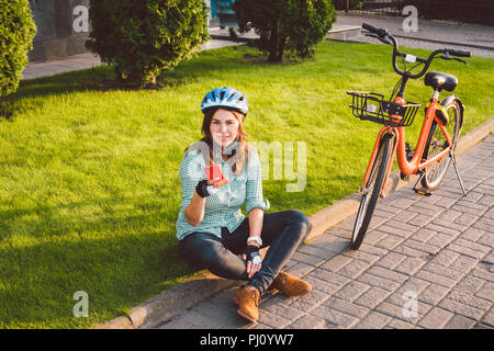 Mensch und Stadt rollenden Fahrrad, umweltfreundliche Verkehrsmittel. Schönen jungen kaukasischen Arbeiterin sitzen ruht auf dem Gras verwendet eine rote mobil Stockfoto