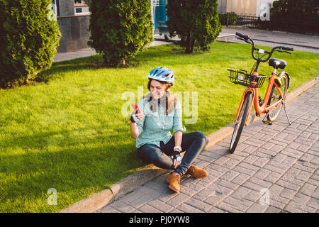 Mensch und Stadt rollenden Fahrrad, umweltfreundliche Verkehrsmittel. Schönen jungen kaukasischen Arbeiterin sitzen ruht auf dem Gras verwendet eine rote mobil Stockfoto