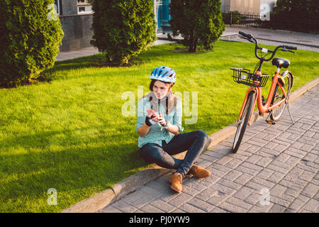 Mensch und Stadt rollenden Fahrrad, umweltfreundliche Verkehrsmittel. Schönen jungen kaukasischen Arbeiterin sitzen ruht auf dem Gras verwendet eine rote mobil Stockfoto