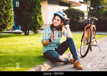 Mensch und Stadt rollenden Fahrrad, umweltfreundliche Verkehrsmittel. Schönen jungen kaukasischen Arbeiterin sitzen ruht auf dem Gras verwendet eine rote mobil Stockfoto