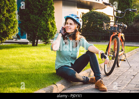 Mensch und Stadt rollenden Fahrrad, umweltfreundliche Verkehrsmittel. Schönen jungen kaukasischen Arbeiterin sitzen ruht auf dem Gras verwendet eine rote mobil Stockfoto