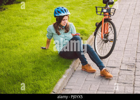 Mensch und Stadt rollenden Fahrrad, umweltfreundliche Verkehrsmittel. Schönen jungen kaukasischen Arbeiterin sitzen ruht auf dem Gras verwendet eine rote mobil Stockfoto