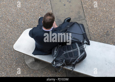 Ein Geschäftsmann auf einem Sitz in der Londoner City sitzen Kontrolle sein Smartphone oder ein mobiles Gerät in Anzug mit Koffer und Taschen von seiner Seite. Stockfoto