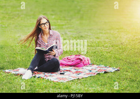 Attraktive junge Frau - Student in Plaid Shirt Brille sitzen und Ihr Lieblingsbuch lesen auf einer grünen Wiese in einen schönen Sommertag. Consept der Erholung, der Bildung und Studium, Neugier, Freizeit Stockfoto