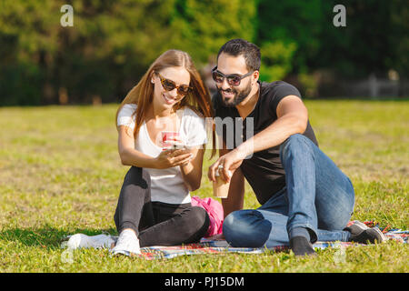 Ziemlich student-Mädchen im weißen T-Shirt mit Sonnenbrille und multikulturellen Studenten - Junge in schwarzen T-Shirt und Sonnenbrille auf der Wiese sitzen, Kaffee trinken und sprechen miteinander Stockfoto