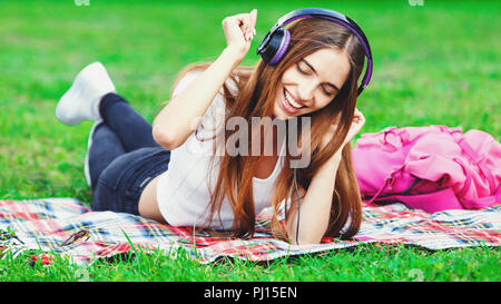 Junge schöne Frau im weißen T-Shirt Musik im Freien auf Kopfhörer liegen auf Gras und suchen gerne Stockfoto