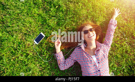 Porträt der schönen europäischen Mädchen in Plaid Shirt Tragen von Kopfhörern Festlegung auf grünem Gras im Park können Sie über Smart phone-Technologie und Kopfhörer im Freien, erholsamen und lächelnd Stockfoto