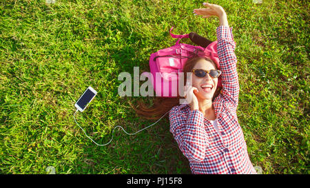 Erstaunlich lange Haare jugendlich Mädchen im karierten Hemd und Sonnenbrille auf ihr rosa Rucksack auf dem Gras und hören Ihre Lieblingsmusik Stockfoto