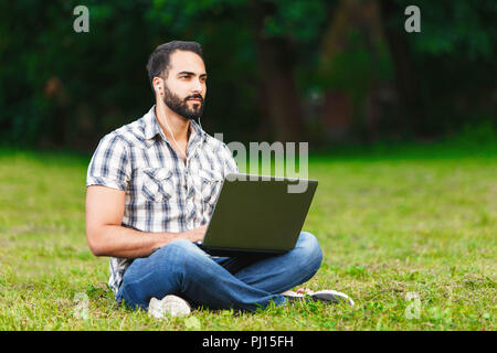 Junge Bartgeier dunkle Haare Mann mit seinem Laptop und das Sitzen auf dem Gras Stockfoto
