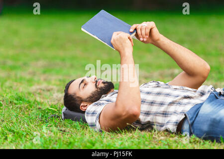 Junge Bartgeier dunkle Haare Mann in Plaid Shirt Verlegung auf dem seinen Rucksack auf dem Gras und sein Lieblingsbuch lesen Stockfoto