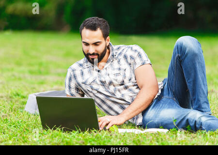 Attraktive schwarze Haare, bärtigen Mann in Plaid Shirt Verlegung auf dem Gras und der Arbeit an seinem Laptop Stockfoto
