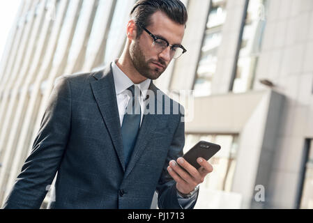 Gut aussehender junger Mann, der in einen kompletten Anzug mit seinem Smart Phone beim Stehen im Freien Stockfoto