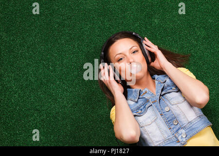 Fröhliche junge Frau mit Kopfhörer genießt in der Musik und blasen Blase im Liegen auf Gras Stockfoto