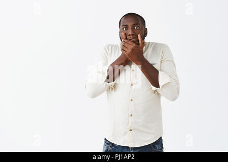 Studio geschossen von Stattlichen nervös african american man Gefühl Bedauern sagen dummer Worte, entsetzt und fassungslos über den Mund mit Palmen und an der Kamera den Blick über graue Wand besorgt Stockfoto
