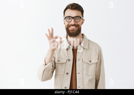 Verlassen Sie sich auf mich, haben Sie alles unter Kontrolle. Freundlich versichern gut aussehenden Freund mit langen Bart in transparenten Gläser, beige Jacke breit grinsend mit glücklich aus, okay oder hervorragende Zeichen Stockfoto