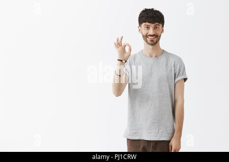 Freund hat alles unter Kontrolle, perfekte unterzeichnen. Glücklich, selbstbewusst und gut aussehender bärtiger Mann im grauen T-Shirt, "Ok" oder "Okay unterzeichnen und breit, die eine exzellente Marke Produkt lächelnd Stockfoto