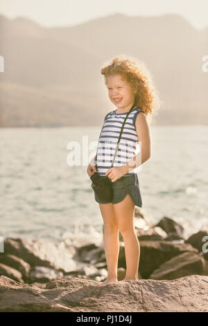 Kleines Mädchen mit Fernglas, sitzt auf einem Felsen in der Nähe eines Meeres Stockfoto