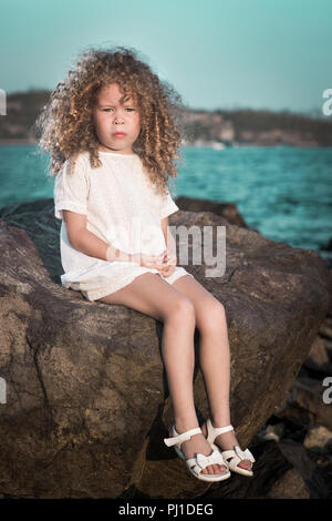 Kleine Mädchen in weißem Kleid auf einem Felsen in der Nähe eines Meeres Stockfoto